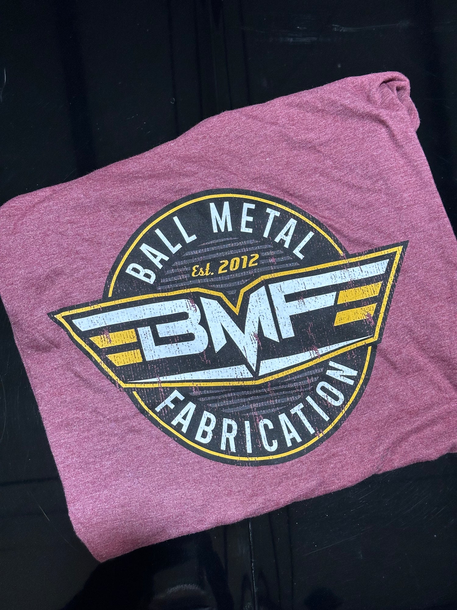 Folded rose color t-shirt with 'Ball Metal Fabrication' logo on a black background